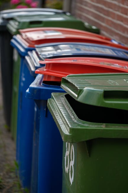 A row of large outdoor wheelie bins in various colors, including green, blue, red, and black, lined up along a paved pathway next to a brick wall. The bins are used for different types of garden waste